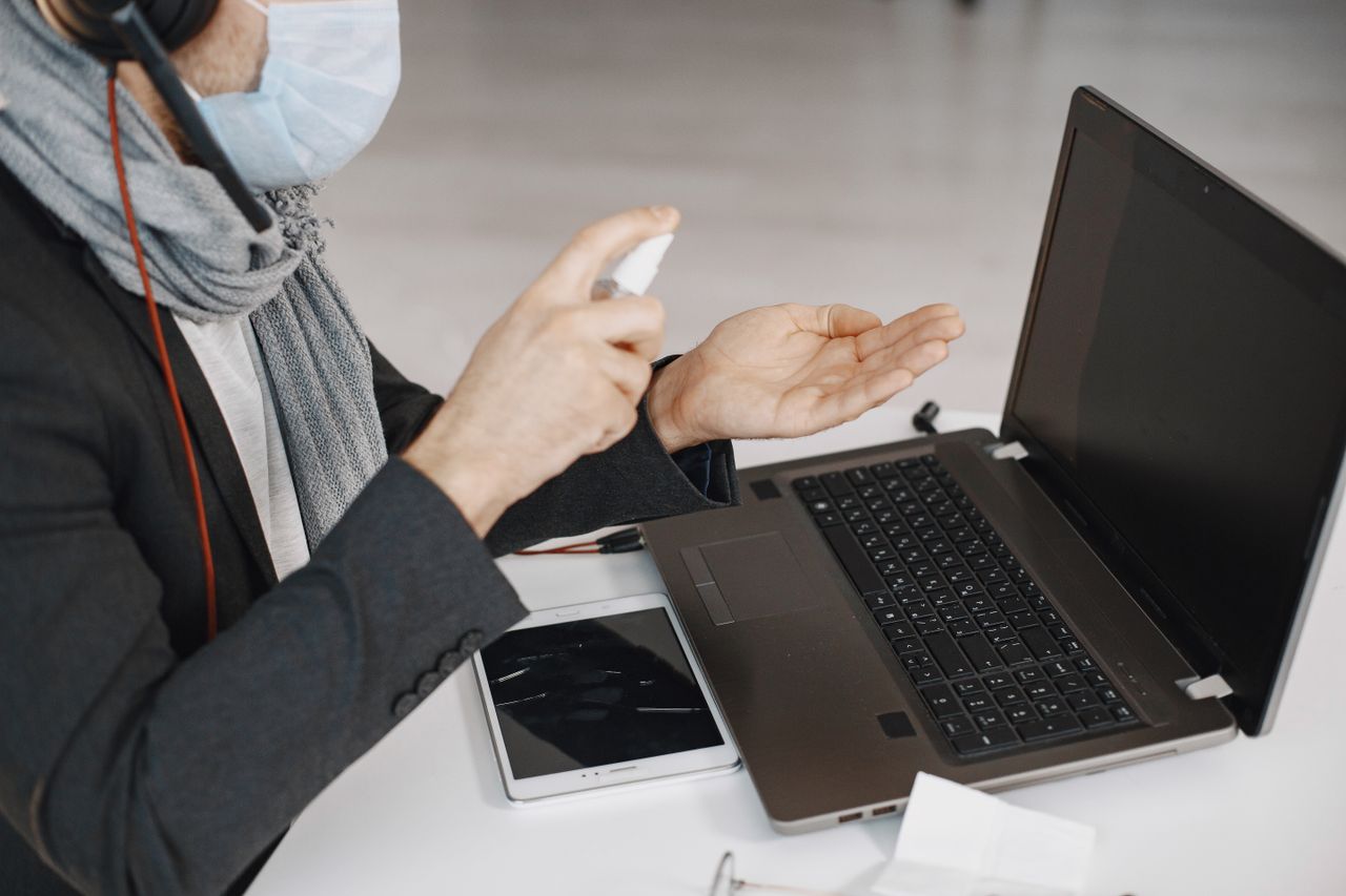 A masked man disinfecting his hands while working on a laptop indoors.