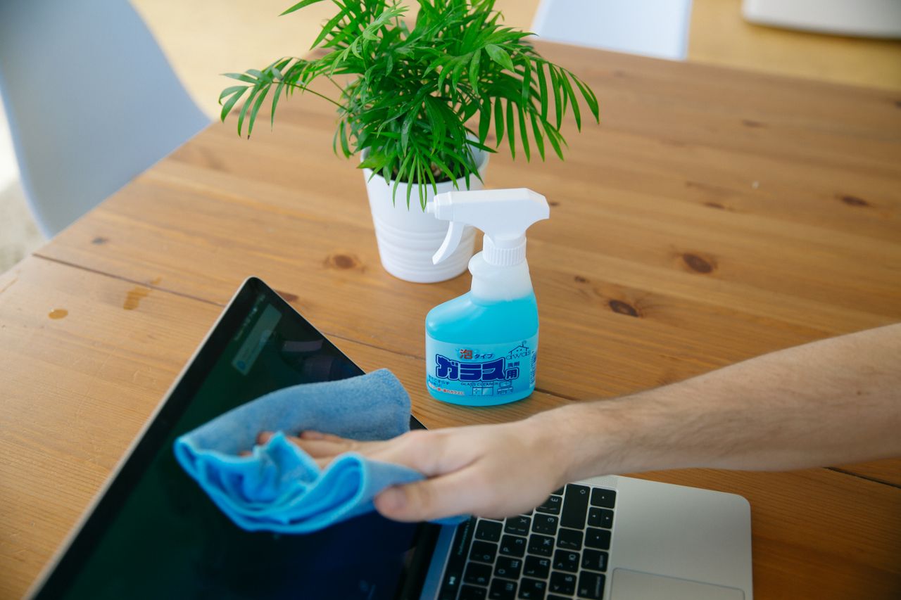 A close-up view of a hand wiping a laptop display with a blue cloth beside a spray bottle on a wooden desk.